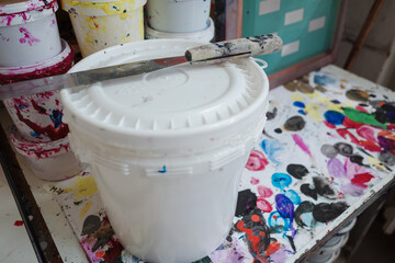 A close-up high-angle view of a large white plastic paint bucket sitting on a workbench covered in colorful paint splatters. A used metal palette knife rests on the bucket's lid.