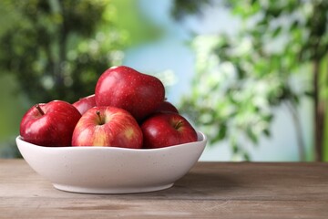 Ripe red apples in bowl on wooden table against blurred green background, closeup. Space for text