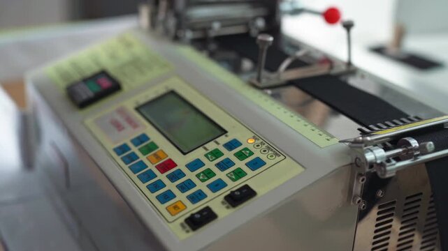 Close-up label printer control panel showing colorful keypad and finger pressing buttons. Small LCD display, paper feed roller, metal clamp, technician