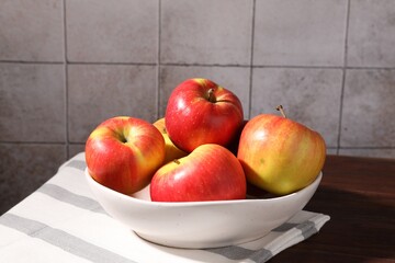 Ripe red apples in bowl on wooden table near grey tiled wall, closeup