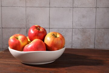 Ripe red apples in bowl on wooden table near grey tiled wall, closeup. Space for text