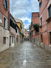 Fototapeta premium A moody, straight-on view down a narrow Venetian street on a wet day. Historic, multi-colored buildings with aged facades and traditional shutters line the wet cobblestone pavement