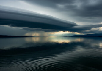 Ominous storm clouds gather over a tranquil lake as dramatic sunbeams break through the dark sky, casting a perfect reflection on the rippling water