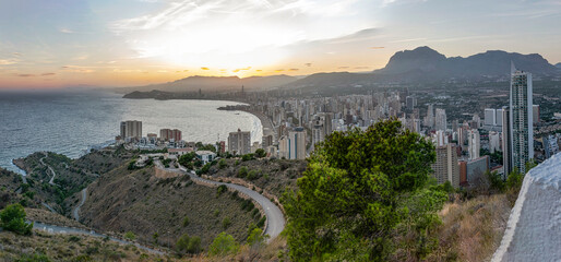 Aerial view from La Cruz de Benidorm coastal city at sunset, showing curving roads, shorelines of Levante and Poniente Beach, tall buildings, distant mountains and calm sea under warm fading sunlight.