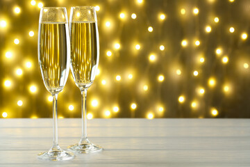 Merry Christmas and Happy New Year. Flute glasses with sparkling wine on white wooden table against golden background with blurred lights, closeup. Space for text