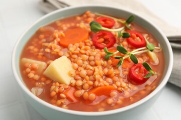 Delicious lentil soup with vegetables in bowl on white tiled table, closeup