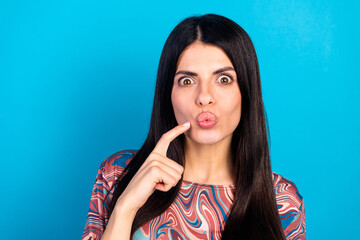 Fototapeta premium Playful young woman with puckered lips pointing to her mouth against a bright blue background wearing a colorful printed shirt