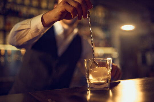 Bartender stirring large ice cube in glass with long bar spoon. Concept of mixology precision, cocktail building, chilled drink preparation, and refined beverage craftsmanship.
