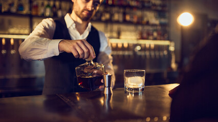 Bartender pouring dark spirit from bottle into jigger beside iced glass. Concept of bar preparation flow, precise drink making, premium serving steps, and mixology accuracy.