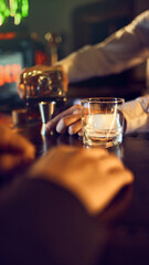Drink with large ice cube on bar counter as bartender prepares bottle nearby. Concept of pre-pour setup, drink assembly workflow, curated bar service, and premium beverage steps.