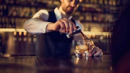 Bartender pouring spirit from jigger into iced glass on polished bar counter. Concept of step-by-step mixology, controlled pour technique, crafted drink quality, and bar workflow.