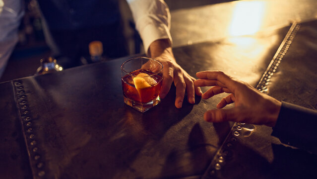 Bartender sliding finished dark drink with citrus peel toward customer. Concept of final service moment, handoff workflow, crafted beverage delivery, and bar hospitality routine.