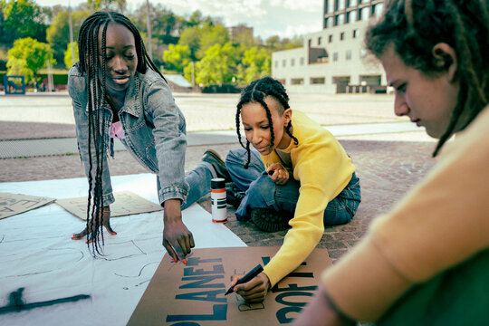 Multicultural young activists preparing a climate protest banner outdoors, painting messages together during an environmental demonstration gathering.	