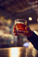 Cocktail with rich amber color held high against blurred bar interior. Concept of premium drink enjoyment, celebratory gestures, crafted spirits, and warm beverage atmosphere.