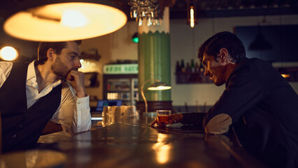 Bartender talking with customer holding drink at bar table. Concept of hospitality service, relaxed after-work conversations, beverage choice guidance, and warm bar ambiance.