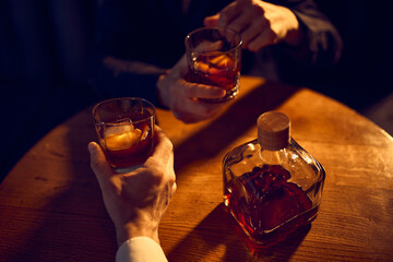 Hands holding whiskey glasses above bottle on wooden table. Concept of bar menu visuals, drink promotion, premium spirits advertising, and lifestyle content for beverage campaigns.