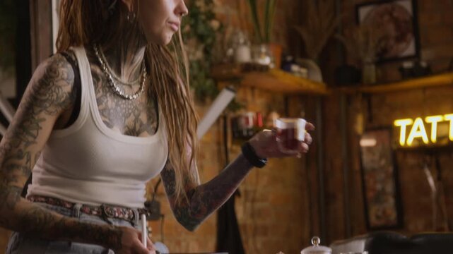Young Caucasian female tattoo artist with dreadlock hairstyle drawing digital sketch on graphic tablet using stylus and enjoying tea break while working in tattoo shop with cozy dim lighting