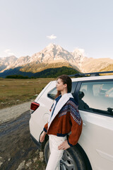 Fototapeta premium Woman, car, mountains, travel, road, nature: Young woman leaning on a white SUV at the roadside with panoramic alpine peaks, cozy jacket, contemplative pose and outdoor adventure mood.
