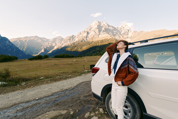 Fototapeta premium Woman by car at roadside with mountains in background, enjoying travel and nature during a scenic road trip, relaxed pose leaning on white SUV at sunset landscape.