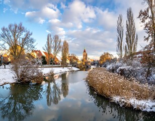 Calm river reflects snow-dusted trees and a quaint town's architecture under a sunny, cloudy sky
