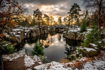 Obraz premium A serene landscape featuring a snowy scene with rocky formations, trees, and a reflective body of water under a dramatic sky at sunset.