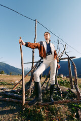 Fototapeta premium Woman standing in mountains wearing boots and jacket, holding a stick in a rural landscape. Outdoor portrait with confident pose, sunlight and scenic field backdrop.