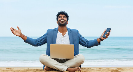 Young Indian businessman sitting cross-legged on the beach with a laptop on his lap and arms open, holding a smartphone