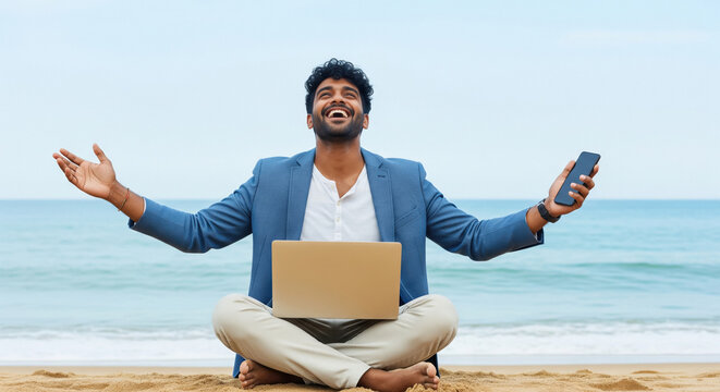 Young Indian businessman sitting cross-legged on the beach with a laptop on his lap and arms open, holding a smartphone