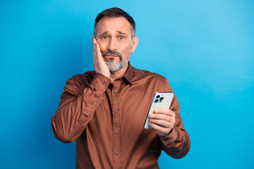 Businessman holds smartphone with concerned expression against blue background ready for a...