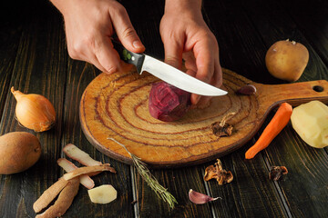 A chef carefully slices a red beet on a wooden cutting board. Surrounding them are various vegetables including onions, carrots, and herbs, ready for preparation in a cozy kitchen