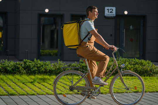 Young man courier wearing a large yellow backpack on his back, riding a classic bicycle on urban street for food delivery service, working in gig economy, daily commuting