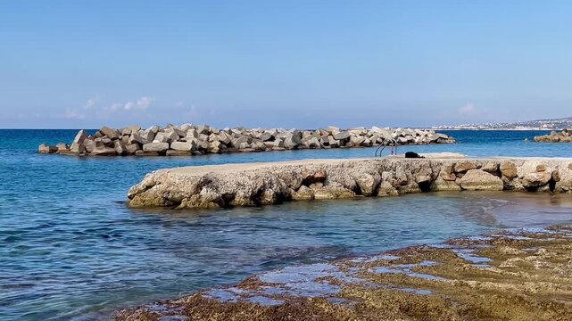 Rocky shoreline at Alyki Beach with clear shallow water and a weathered concrete pier leading toward protective breakwater blocks, creating a calm Mediterranean coastal scene