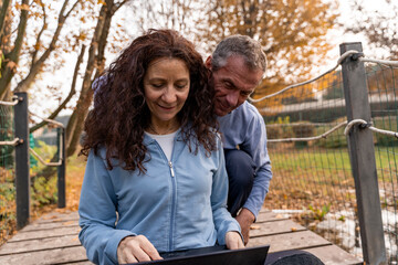 Older couple sitting on a rustic bridge, looking at a laptop together outdoors