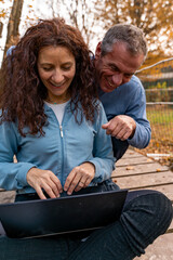 Middle-aged couple laughing and using laptop together on a wooden bridge in nature