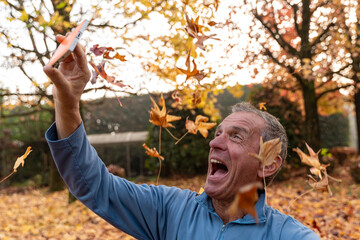 Happy man holding smartphone, recording or taking selfie with falling autumn leaves in park