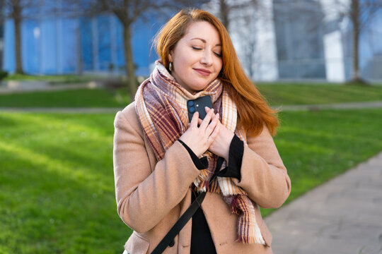 Redhead woman feeling connected and happy, embracing her smartphone with affection in an urban park