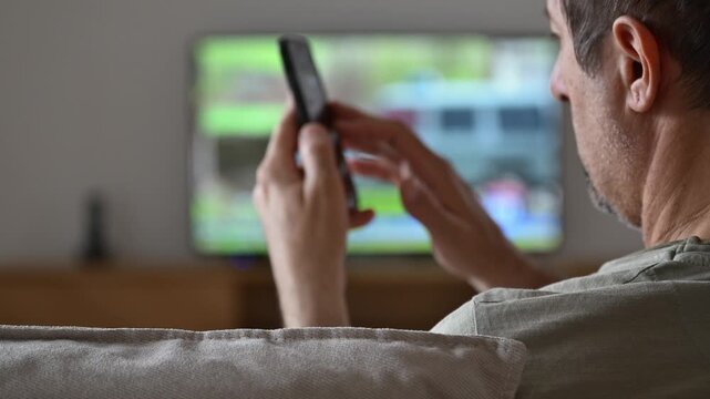 Man using smartphone while watching television at home, multitasking and browsing on mobile device in relaxed living room setting. 4K.