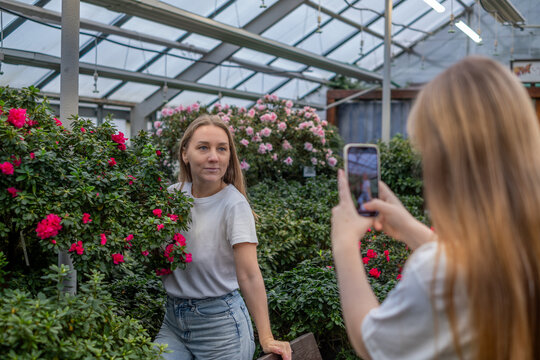 Young woman posing among vibrant flowers in a greenhouse while her teenage daughter captures the moment with a smartphone, showcasing their close family bond and togetherness
