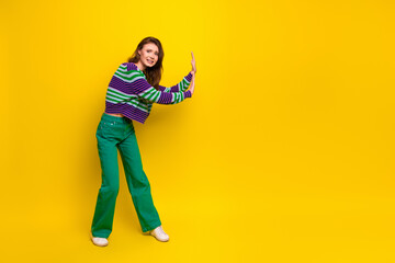 Young woman in striped sweater green pants smiles while press against bright yellow background