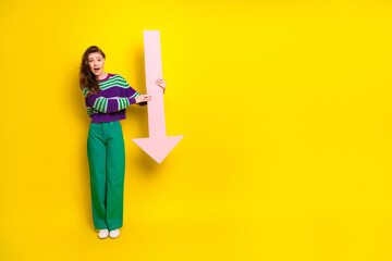 Young woman in striped sweater and green pants holds pink arrow against bright yellow background