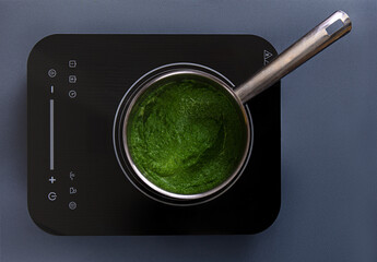 A pot filled with a smooth, bright green sauce sits on an induction cooktop. The scene captures a kitchen environment with modern appliances and natural brightness.