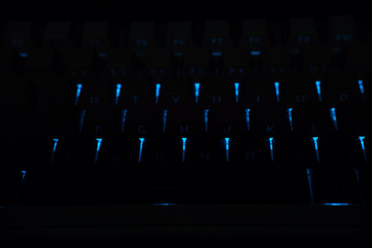 Low-key close-up of a mechanical keyboard in total darkness, illuminated only by vertical streaks of blue LED backlighting. The keycaps are silhouetted against the glow, creating a mysterious hi tech