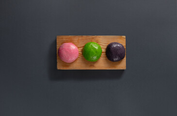 Three round traditional sweets in pink, green, and purple shades are neatly placed on a wooden board. Their vibrant colors attract the eye, adding charm to the simple presentation.