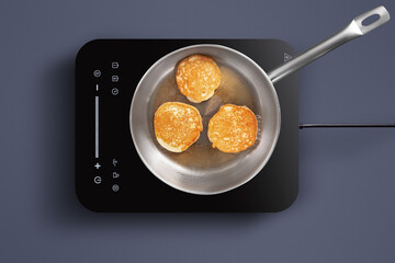 Three golden-brown pancakes are sizzling in a stainless-steel pan on an induction cooktop. The cozy kitchen offers a warm atmosphere, perfect for breakfast preparation.