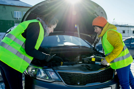 Two women in reflective safety vests working together under open car hood on sunny snowy day