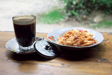 : Glass of black coffee on a wooden table with a metal spoon and lid. Blurred background shows golden fried banana fritters on a vintage enamel plate, depicting a traditional Indonesian rural snack.