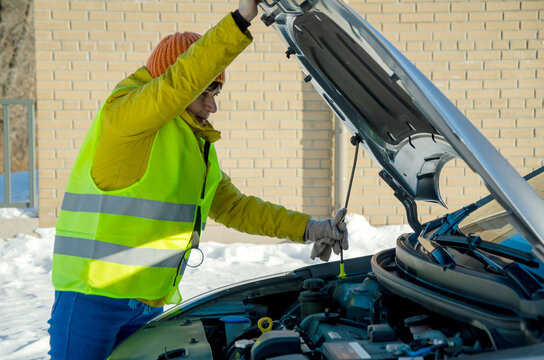Senior woman in reflective safety vest checking car engine under open hood on snowy winter day - Powered by Adobe