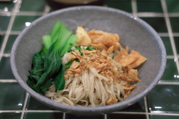 Bowl of dry noodles topped with fried wonton crisps, blanched bok choy, fried shallots, and chopped scallions. Served in a grey ceramic bowl on a green square-tiled table. Traditional Asian cuisine.
