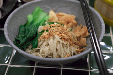 Bowl of dry noodles topped with fried wonton crisps, blanched bok choy, fried shallots, and chopped scallions. Served in a grey ceramic bowl on a green square-tiled table. Traditional Asian cuisine.