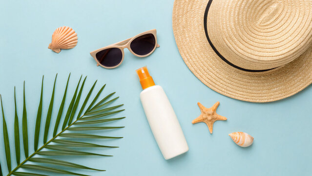 uv-safety awareness month A flat lay arrangement includes sunscreen, dark sunglasses, a straw hat, and seashells on a light blue background, perfect for a sunny beach day.